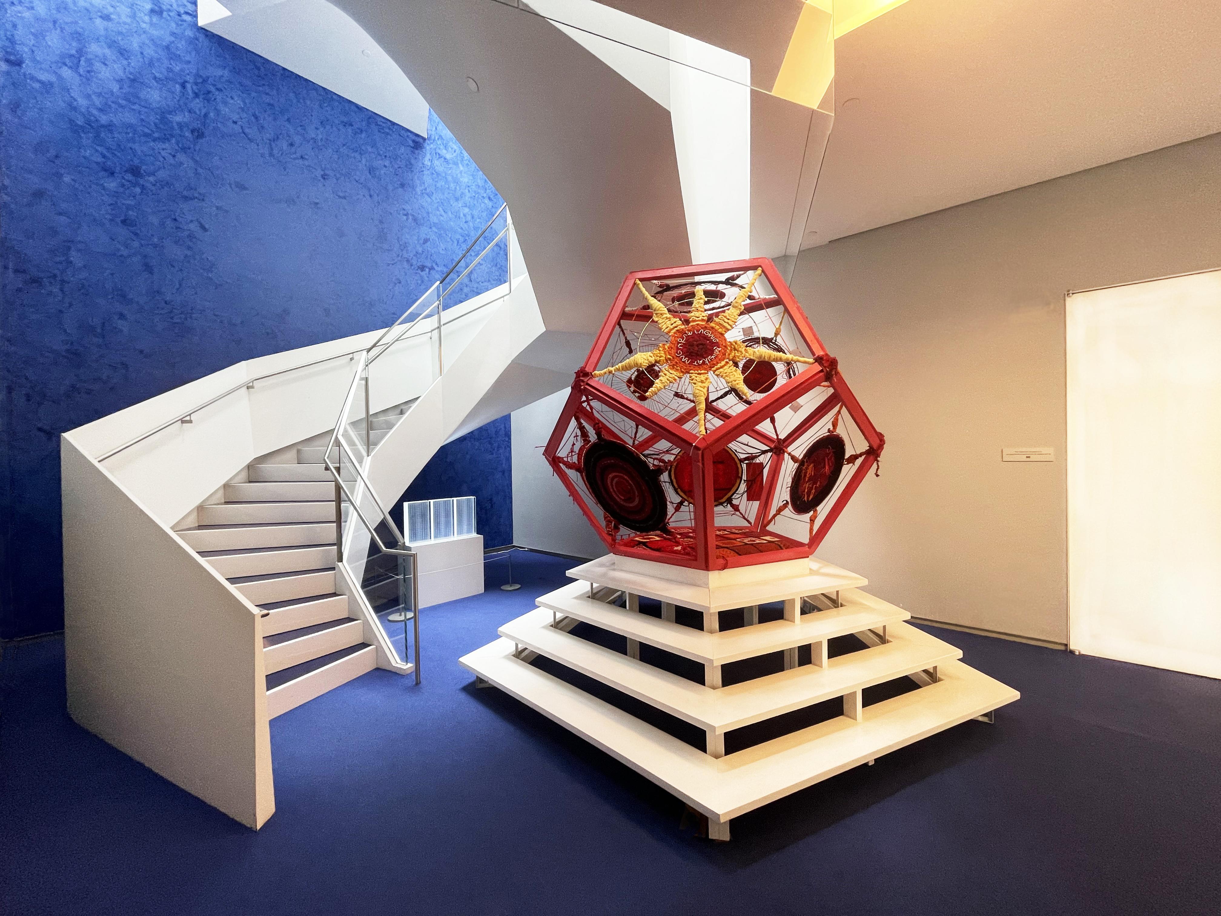 longer view of 3D red pentagon art installation, with sweeping white staircase in the background
