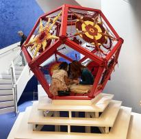 Young children sit inside a 3D red hexagon with designs on it atop a stair pedestal