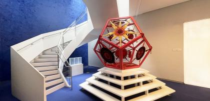 longer view of 3D red pentagon art installation, with sweeping white staircase in the background